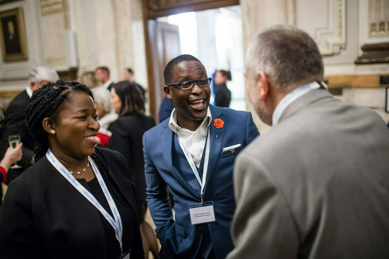 Three people in formal attire are engaged in a lively discussion at an event. They are smiling, wearing name tags, and standing in an ornately decorated room. The man in the blue suit has a lapel pin. Other attendees can be seen in the background. James Gifford-Mead Photography - Event Photographer London