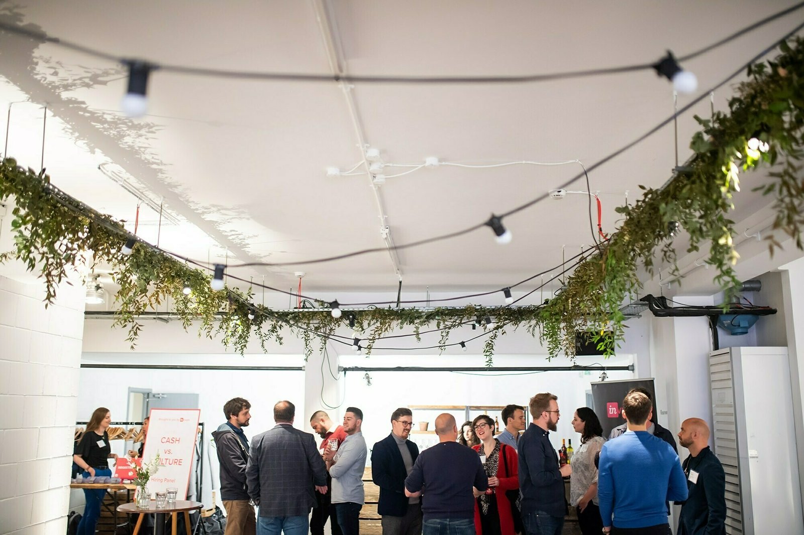 A group of people socializing at an indoor event. The room is decorated with hanging greenery and string lights. Some attendees are gathered near a board. Casual and lively atmosphere