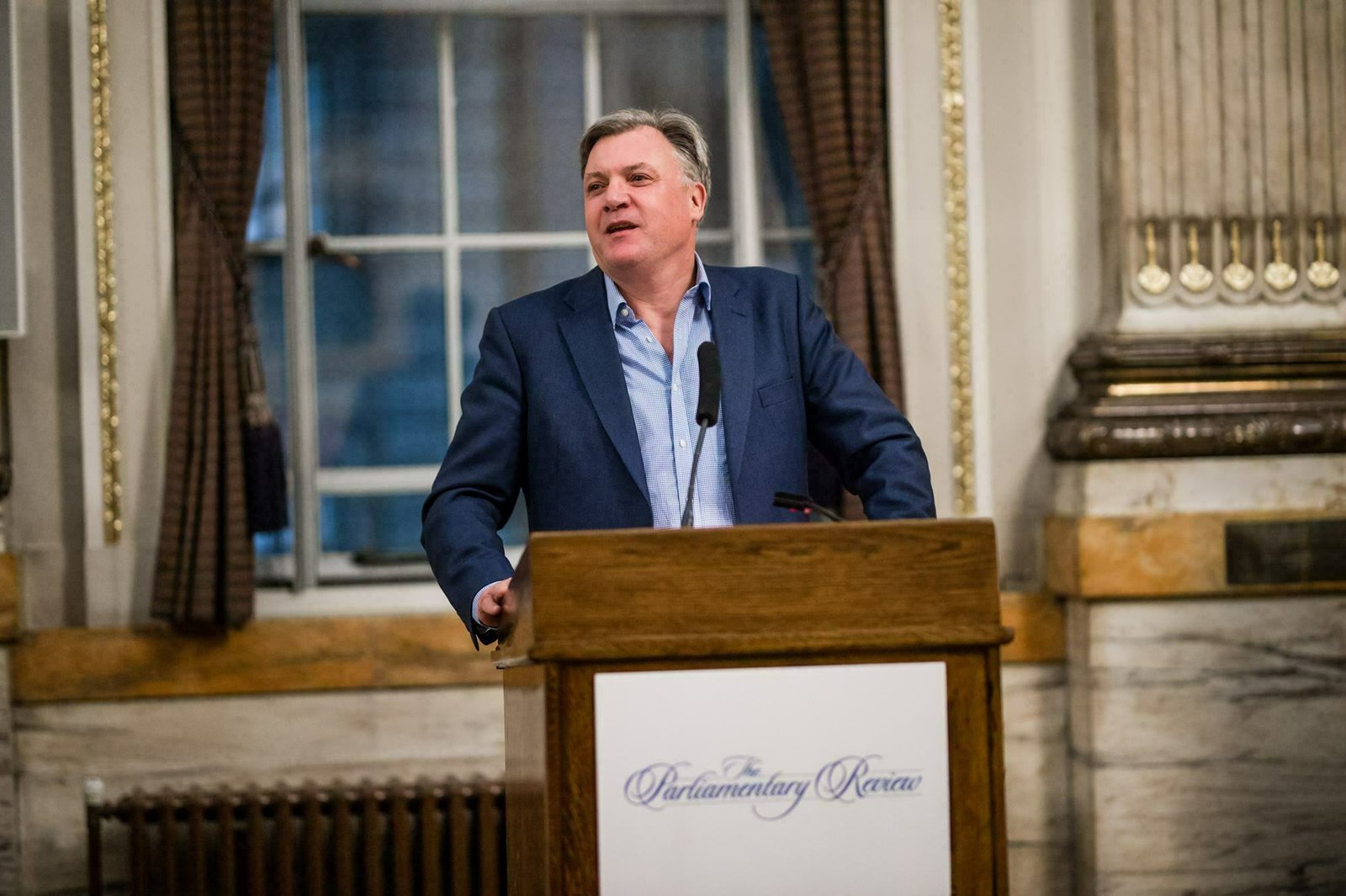 A man in a blue suit speaks at a podium with a microphone. Behind him are large windows with curtains in a grand room featuring marble and decorative columns. The podium displays a sign reading "The Parliamentary Review. James Gifford-Mead Photography - Event Photographer London