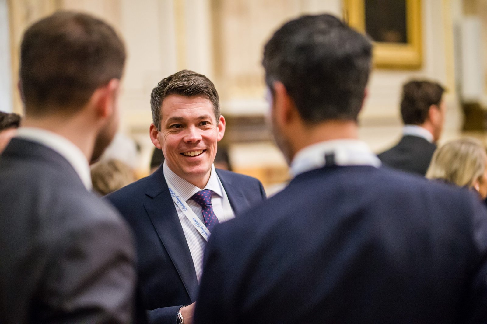 A man in a suit smiles as he converses with two others at a formal gathering. The background is softly focused, suggesting an elegant setting. He appears engaged and attentive amidst a bustling, professional crowd. James Gifford-Mead Photography - Event Photographer London