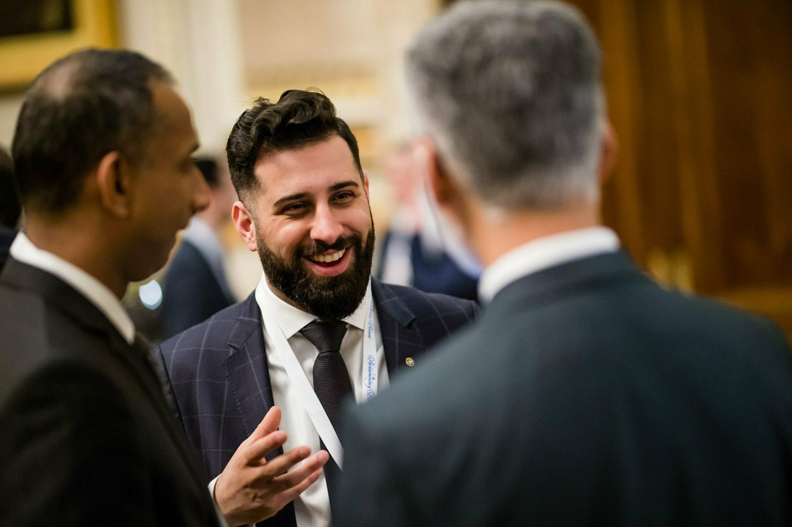 Three men in suits engage in conversation indoors, with one man smiling and gesturing while the others listen. The background is softly blurred, suggesting a formal setting like a conference or networking event. James Gifford-Mead Photography - Event Photographer London