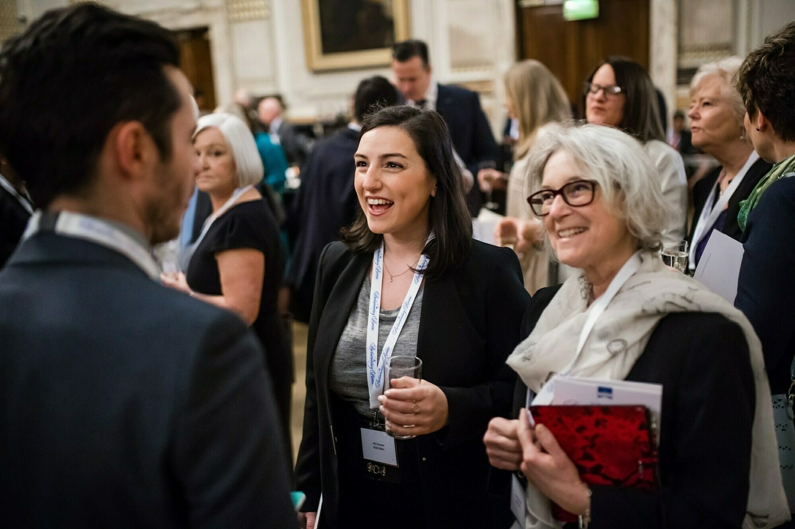 A group of people in business attire are mingling at a formal event. Two women are engaged in conversation with a man, holding drinks and papers. The background shows more attendees in a grand room with ornate decor. James Gifford-Mead Photography - Event Photographer London