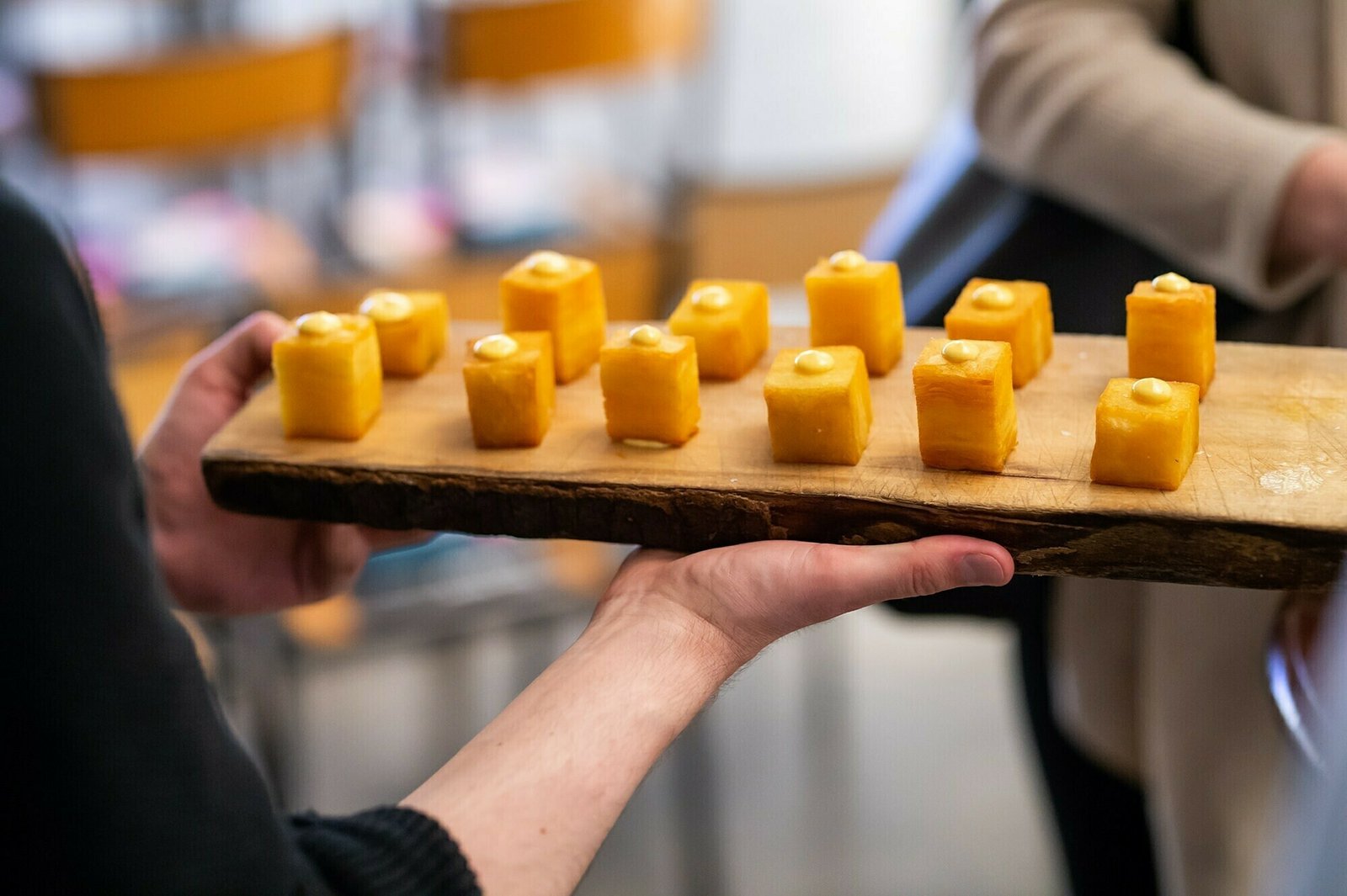 A person holds a wooden board with cubed yellow desserts, each topped with a small dollop of cream. Several blurred chairs are visible in the background. James Gifford-Mead Photography - Event Photographer London