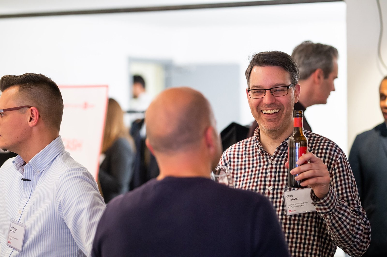 A group of people at a social event, engaging in conversation. A man in a checkered shirt smiles while holding a drink bottle. Others are nearby, chatting and wearing name tags. The setting appears to be informal and friendly. James Gifford-Mead Photography - Event Photographer London