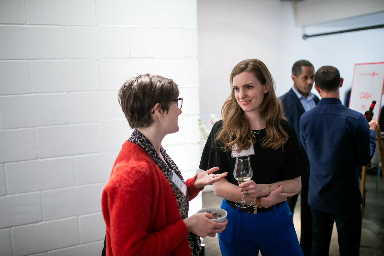 Two people are conversing at a social event. One, wearing a red cardigan and glasses, gestures while holding a bowl. The other, in a black top and blue pants, holds a wine glass. Other attendees are visible in the background. James Gifford-Mead Photography - Event Photographer London