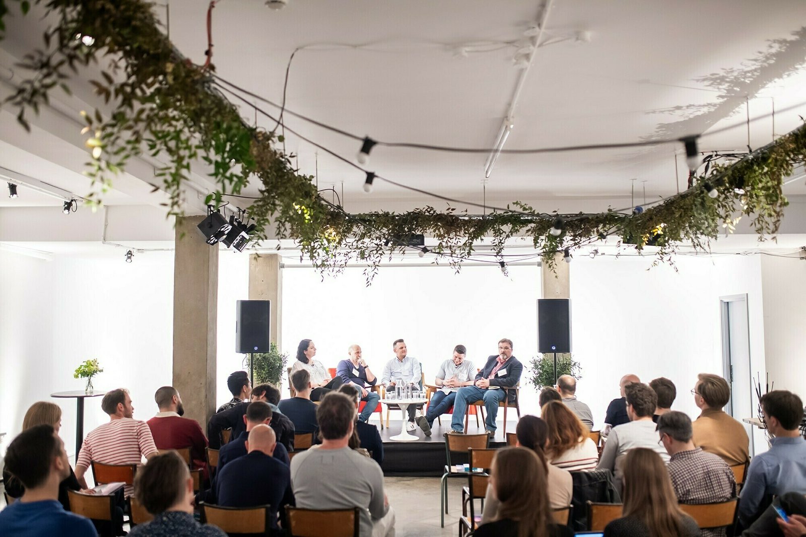 A panel discussion is taking place in a modern conference room with hanging greenery. Four panelists are seated on stage with microphones. An audience sits attentively in rows of chairs, listening. James Gifford-Mead Photography - Event Photographer London