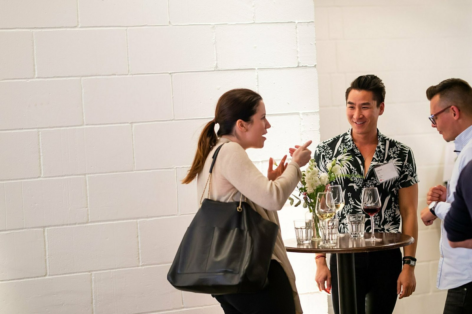 Three people are standing around a small round table, talking and smiling. The table has wine glasses and small flower arrangements. The background features a white brick wall. James Gifford-Mead Photography - Event Photographer London