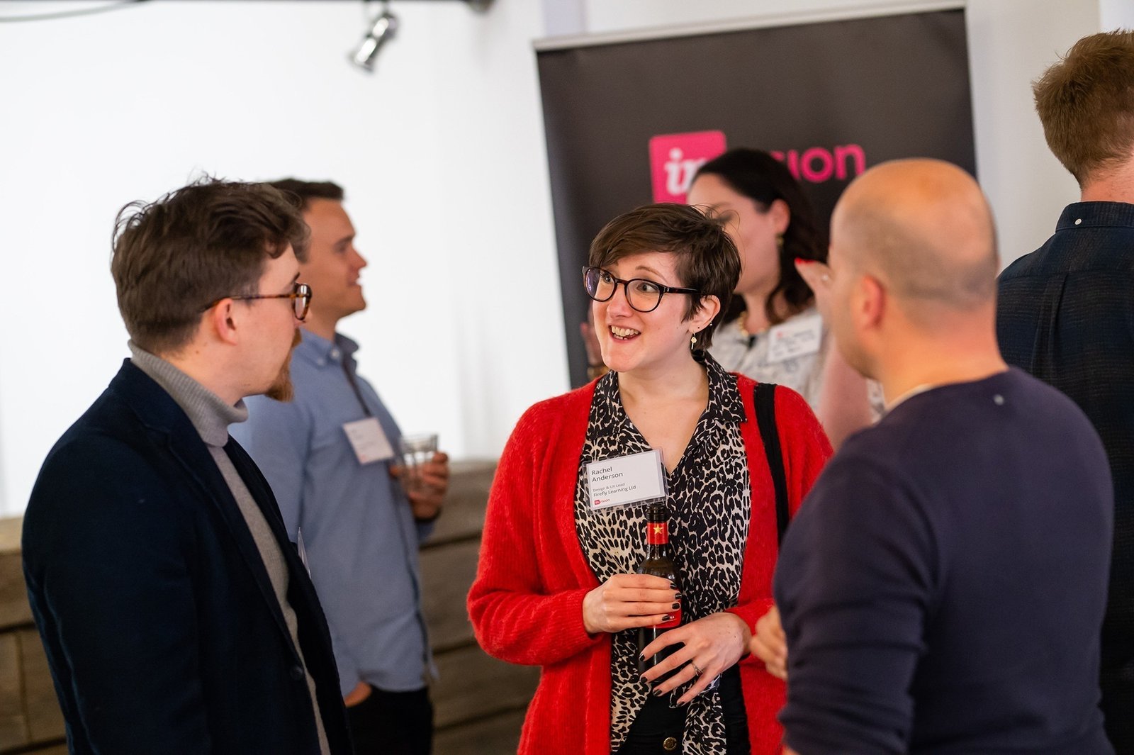 A group of people are engaged in a casual conversation at an indoor event. They are standing, holding drinks, and wearing name tags. A banner in the background is partially visible. One person in a red cardigan is smiling and listening attentively. James Gifford-Mead Photography - Event Photographer London