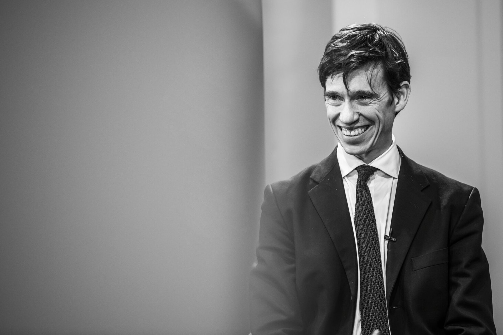 A man in a suit and tie is smiling while sitting in a chair. The image is in black and white, and he is positioned to the right with a blurred background on the left
