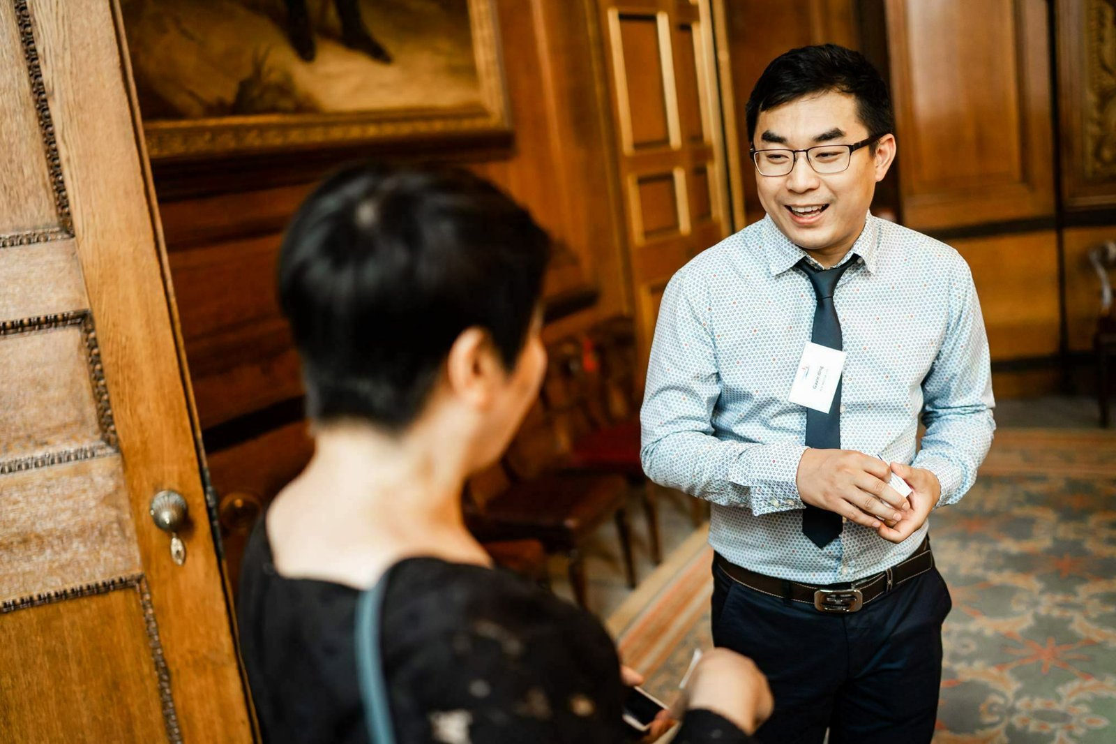 A man in glasses and a patterned shirt with a tie is smiling and talking to a woman with short hair in a dimly lit room with wooden walls and a painting. James Gifford-Mead Photography - Event Photographer London