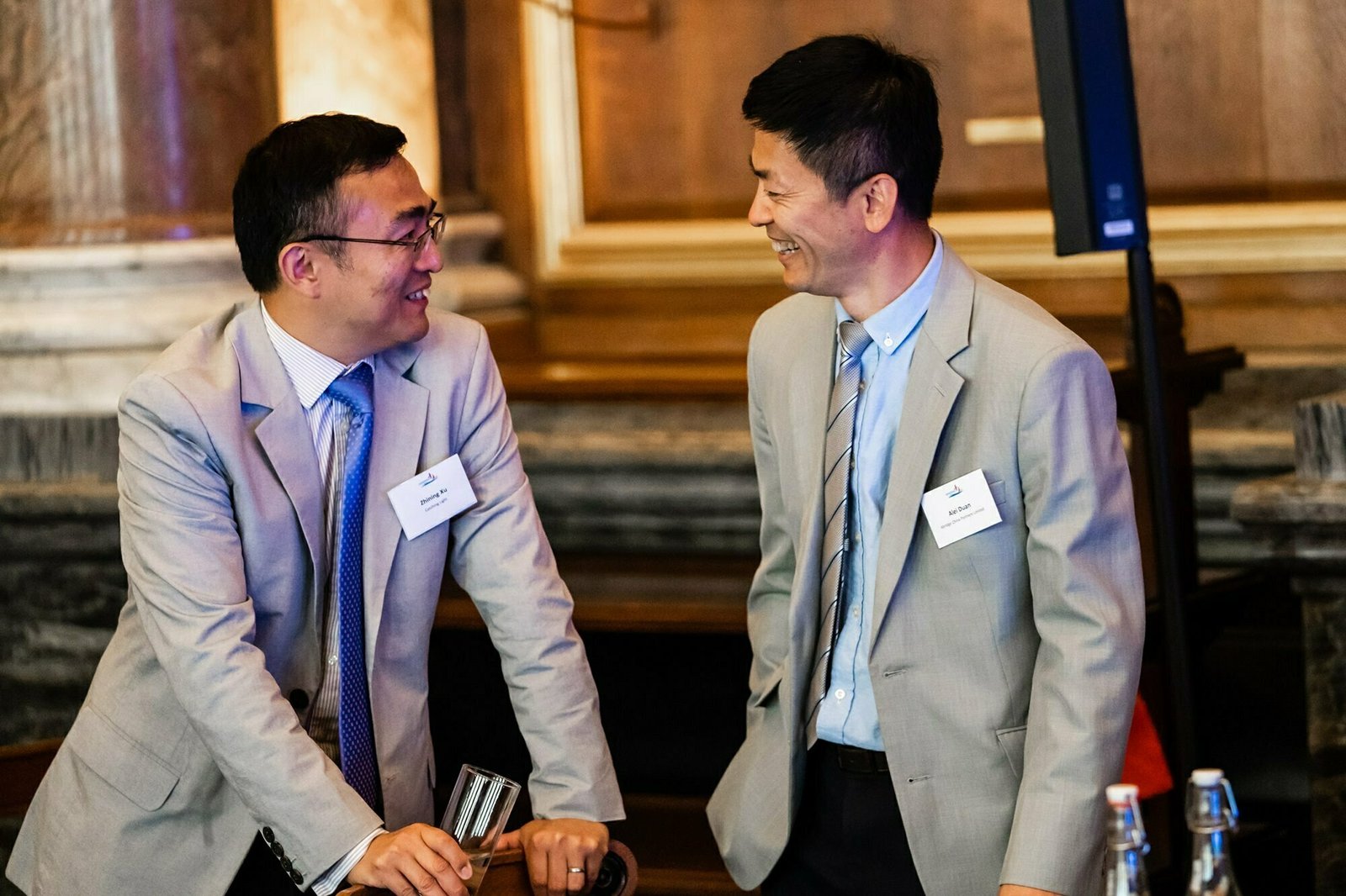 Two men in suits smiling and talking to each other in a formal setting. Both wear glasses and have conference name tags. They stand near a table with bottles and papers. The background features wooden paneling and marble accents. James Gifford-Mead Photography - Event Photographer London