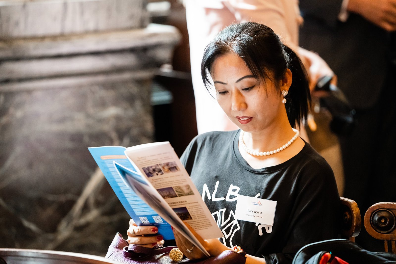 A woman with dark hair, wearing a pearl necklace and a name tag, is seated and reading a booklet. She appears focused and is holding the booklet open. The background is slightly blurred, suggesting a public setting. James Gifford-Mead Photography - Event Photographer London