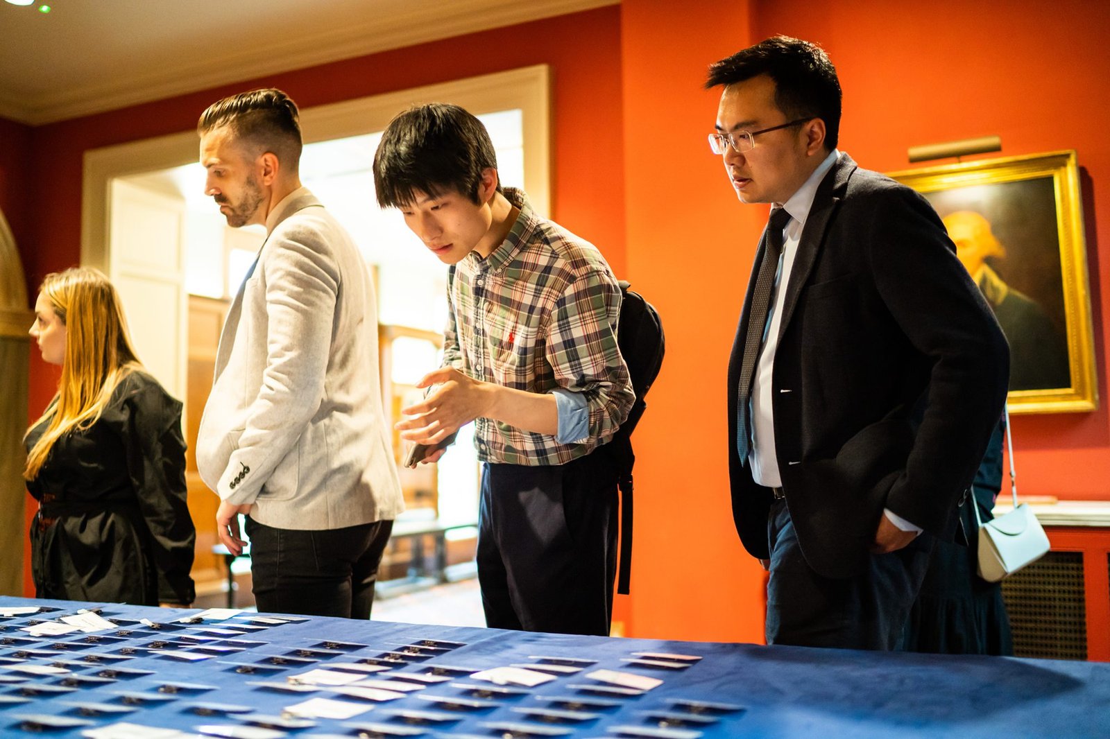 Four people stand around a table covered with rows of small cards. They appear focused, examining and engaging with the cards. The room has red walls and framed art, creating a formal setting. James Gifford-Mead Photography - Event Photographer London