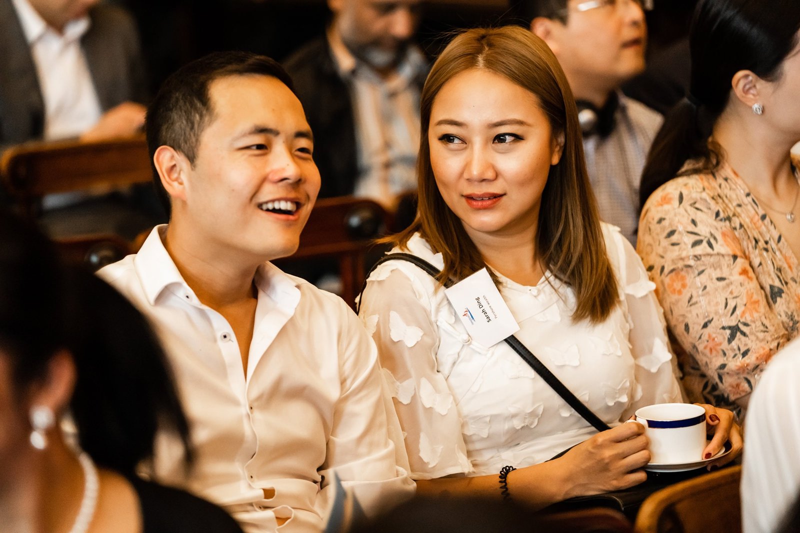Two people seated indoors engage in conversation. The man, wearing a white shirt, is smiling, while the woman beside him, also in white, holds a cup and listens attentively. Other blurred attendees are visible in the background. James Gifford-Mead Photography - Event Photographer London