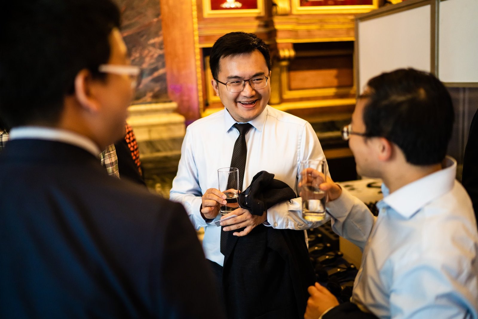 Three people in formal attire are conversing and smiling at a social event. One man is holding a drink and a blazer. They appear to be in a room with ornate decor and a warm ambiance. James Gifford-Mead Photography - Event Photographer London