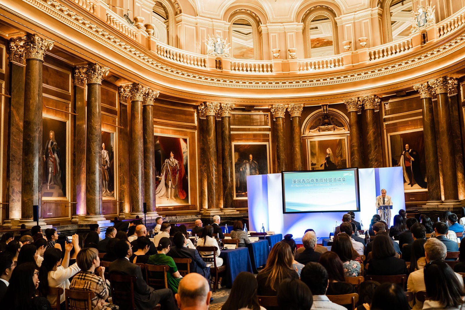 A large group of people seated in an ornate hall with marble columns and framed portraits on the walls. A speaker stands at a podium next to a large screen displaying text in Chinese. The ceiling is dome-shaped with intricate moldings