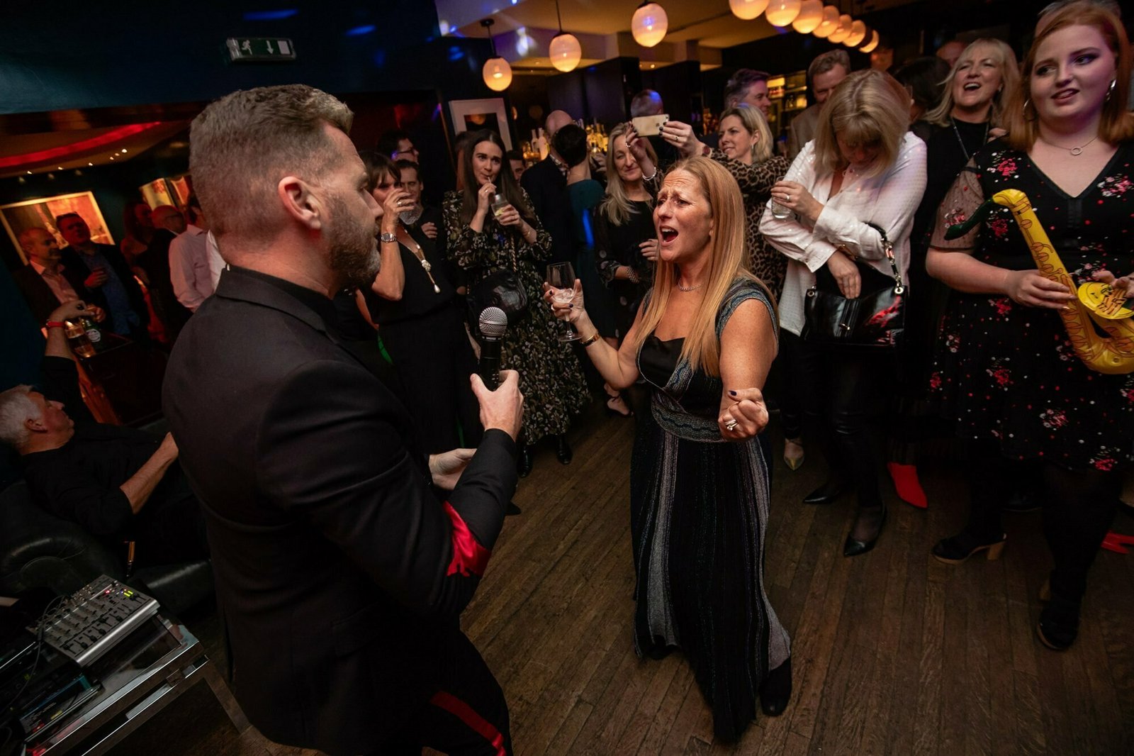 A lively party scene with a man holding a microphone singing to a woman who is enthusiastically dancing and singing along. People are gathered around, smiling and enjoying the performance amid festive, dim lighting and hanging globe decorations. James Gifford-Mead Photography - Event Photographer London