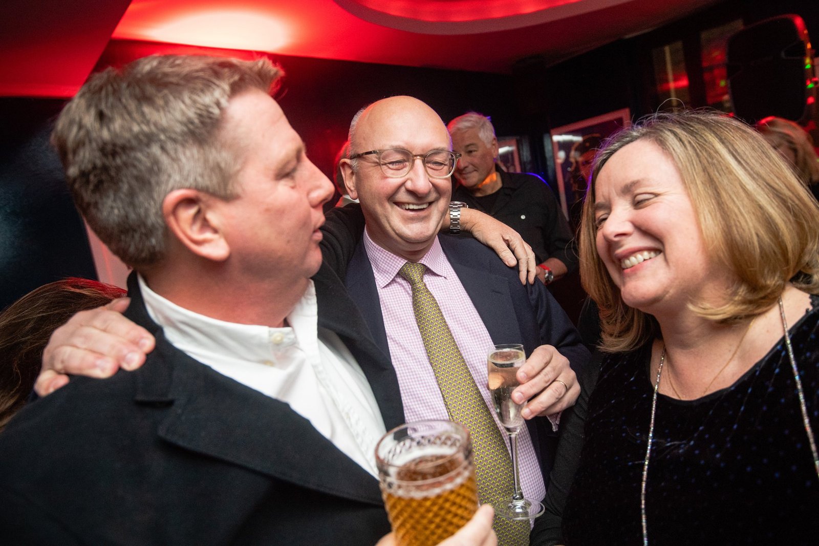 Three people, two men and a woman, are laughing and holding drinks at a social event. They appear to be in a lively atmosphere with red lighting in the background, suggesting a party or celebration. James Gifford-Mead Photography - Event Photographer London
