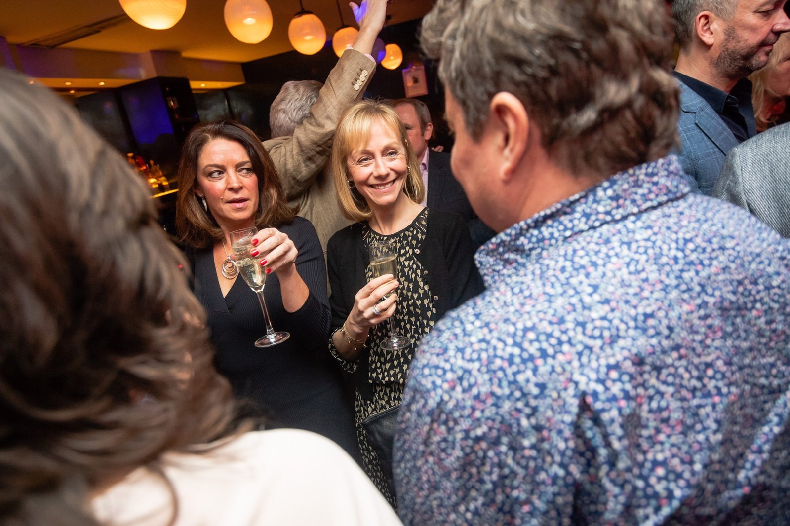 A lively party scene with people holding drinks and chatting in a crowded room. The focus is on two women holding glasses. One woman is wearing a black outfit and smiling, while the other has an animated expression. More guests are mingling in the background