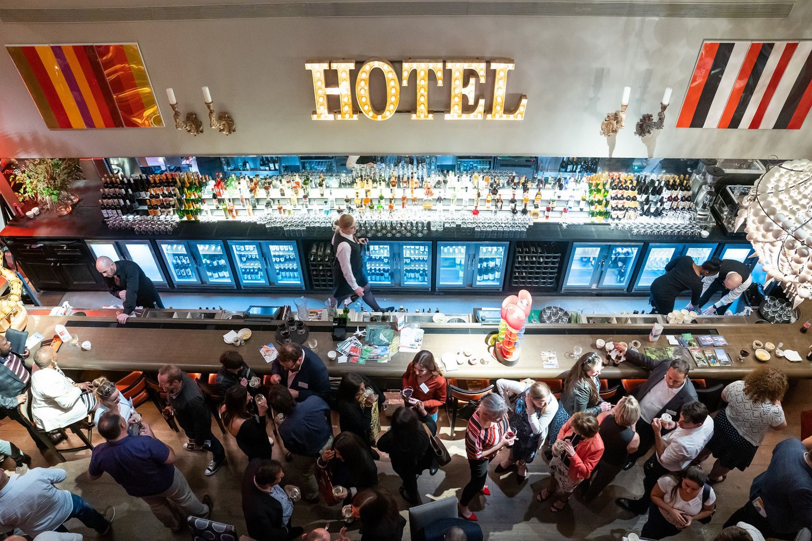 Aerial view of a bustling hotel bar with people socializing. A bartender works behind the counter filled with bottles. A large, illuminated "HOTEL" sign hangs above, and the bar has modern decor with vibrant wall art