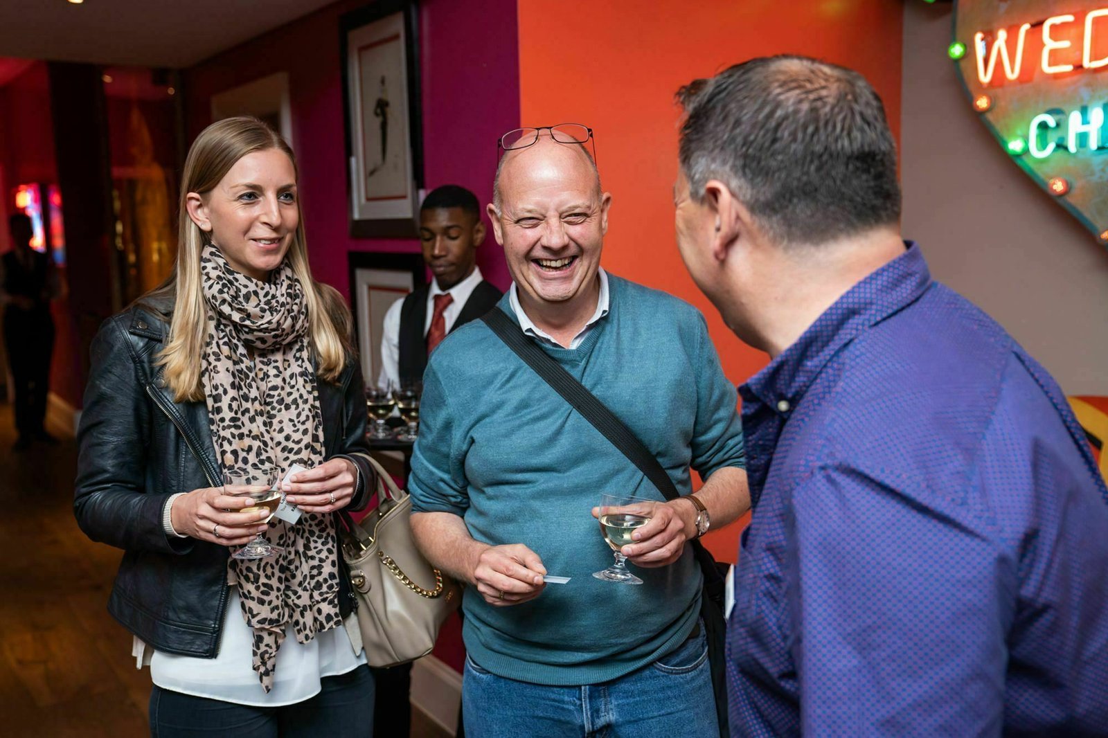 Three people are socializing at an indoor event. A woman in a patterned scarf and a man in a teal sweater are holding drinks, smiling, and chatting with a man in a blue shirt. A server with a tray is visible in the background. James Gifford-Mead Photography - Event Photographer London