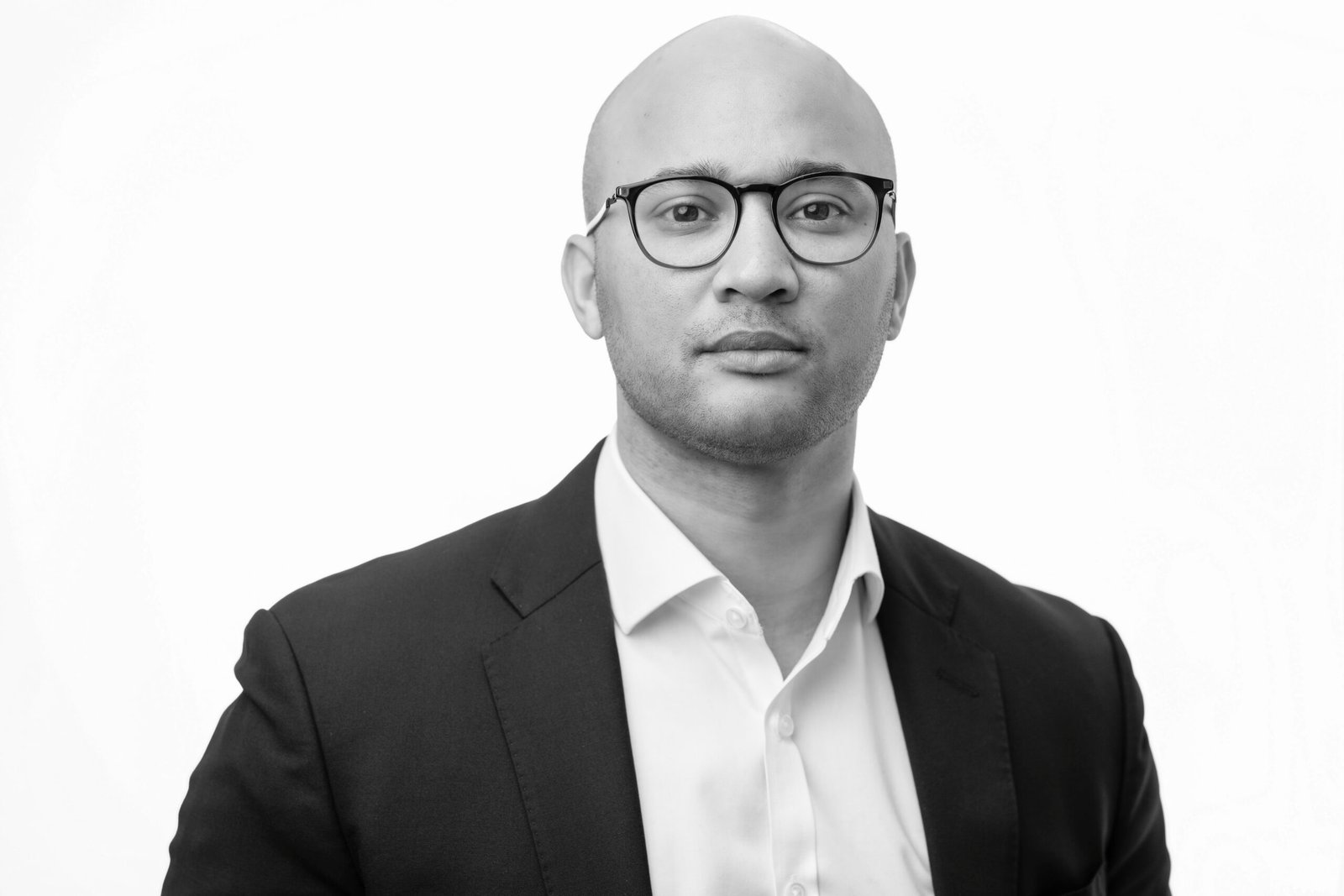 A person in a black suit and white shirt is looking straight ahead. They are wearing glasses and have a neutral expression. The background is white. The image is in black and white
