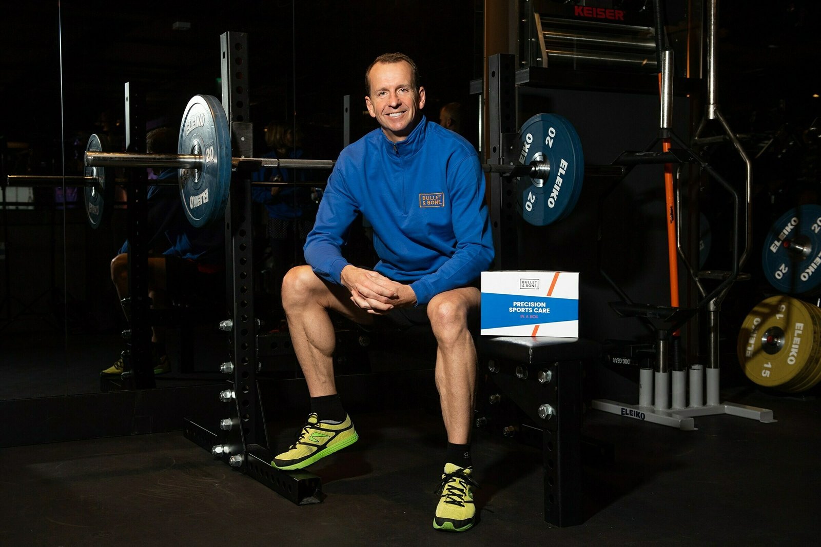 A person wearing a blue sports jacket and neon green sneakers sits on a weight bench in a gym. There are weights on a rack behind them, and a sign that reads "Precision Sports Care" rests nearby. They are smiling and looking at the camera