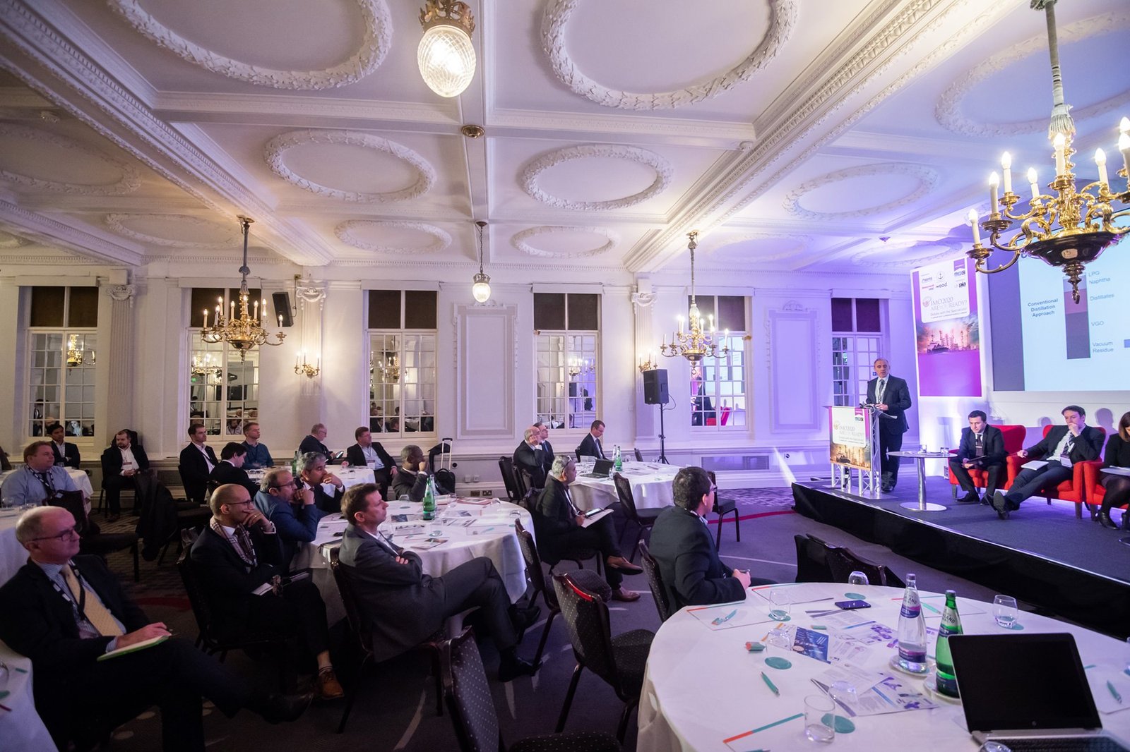 A conference room with people seated at round tables, listening to a speaker at a podium. The room has ornate ceilings, chandeliers, and large windows. A projection screen shows a presentation slide