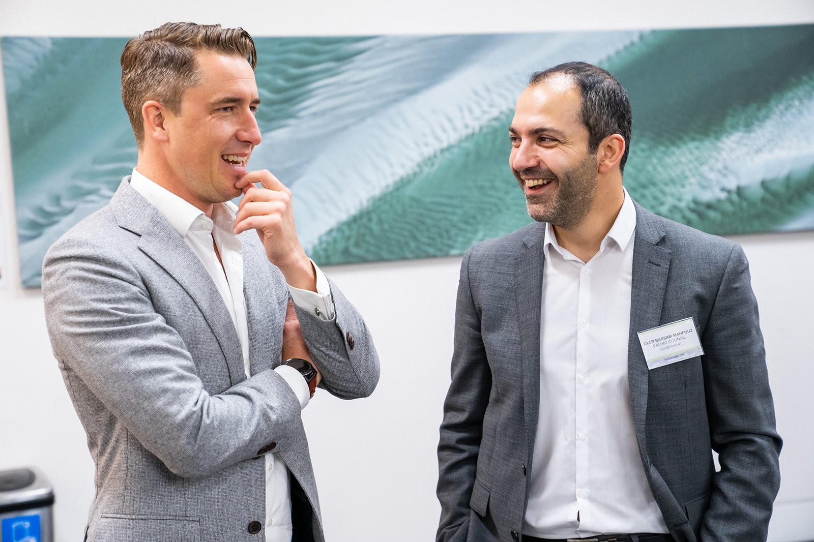 Two men in suits are engaged in conversation. One man is touching his chin while the other is smiling. They are standing in front of a green and white artwork. Both are wearing name badges. James Gifford-Mead Photography - Event Photographer London