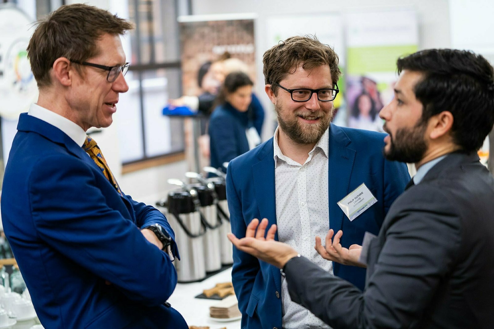Three men in suits engage in a lively conversation at an indoor event. One man gestures while talking, and the others listen with interest. A table with coffee dispensers and cups is visible in the background. James Gifford-Mead Photography - Event Photographer London