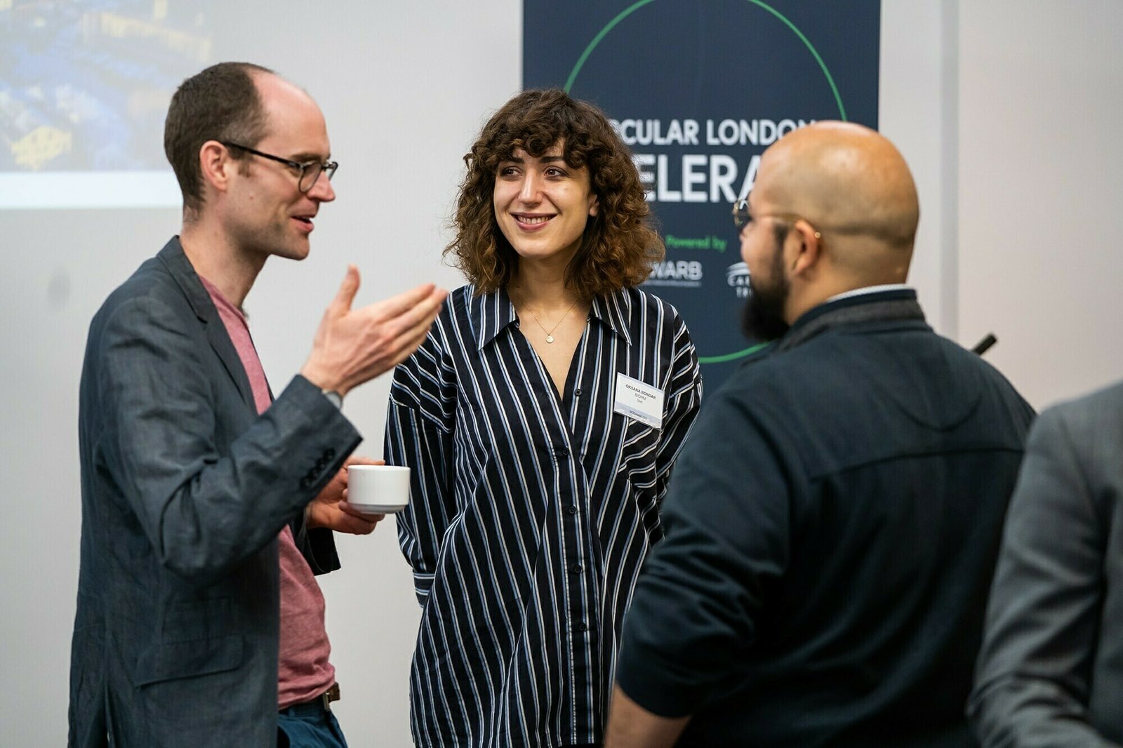 Three people are engaged in conversation at an event. One person holds a coffee cup while speaking. A sign with partially visible text is in the background. The setting appears to be professional or conference-related. James Gifford-Mead Photography - Event Photographer London