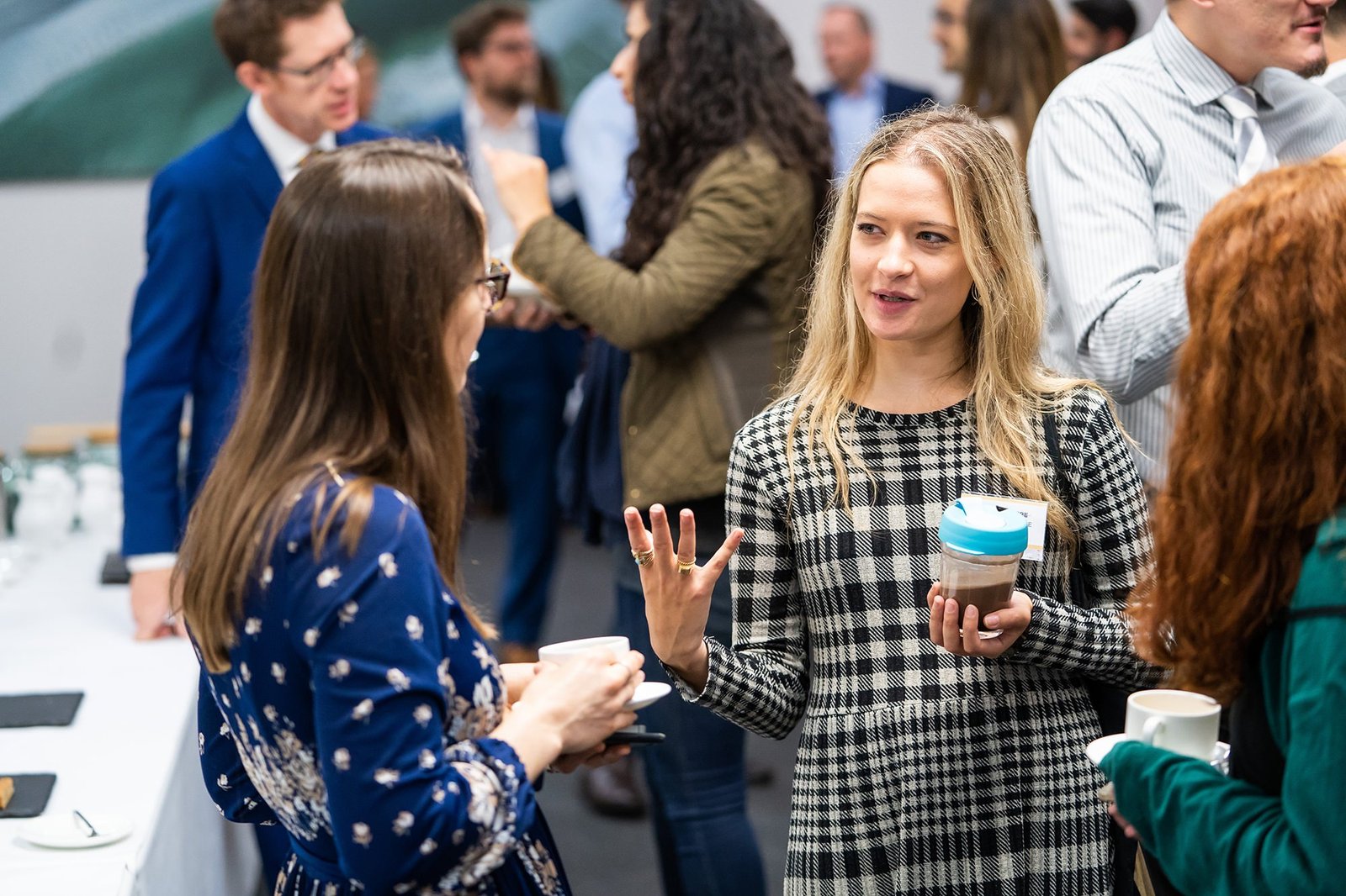 A group of people socialize at a networking event. Two women in the foreground engage in conversation, holding cups. The setting is informal, and others mingle in the background, some holding drinks. James Gifford-Mead Photography - Event Photographer London