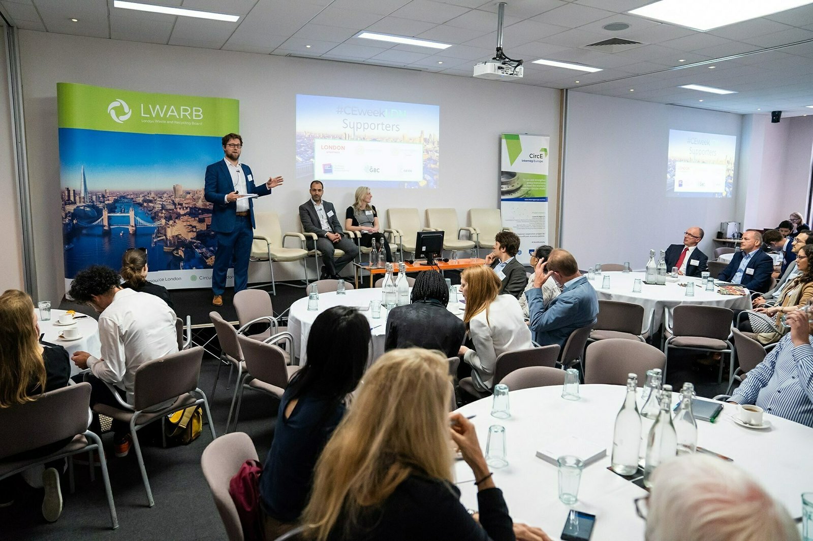 A man stands beside a screen during a presentation in a conference room. Two people are seated on stage. Attendees sit at round tables facing the stage. Banners and a large window are in the background. James Gifford-Mead Photography - Event Photographer London
