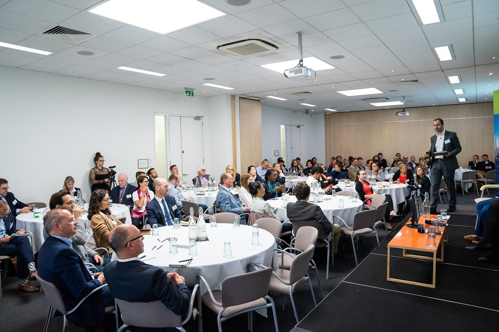 A conference room filled with people seated at round tables. A speaker stands on a stage in front of the audience. The room is brightly lit with ceiling lights, and there's a projector mounted on the ceiling. James Gifford-Mead Photography - Event Photographer London
