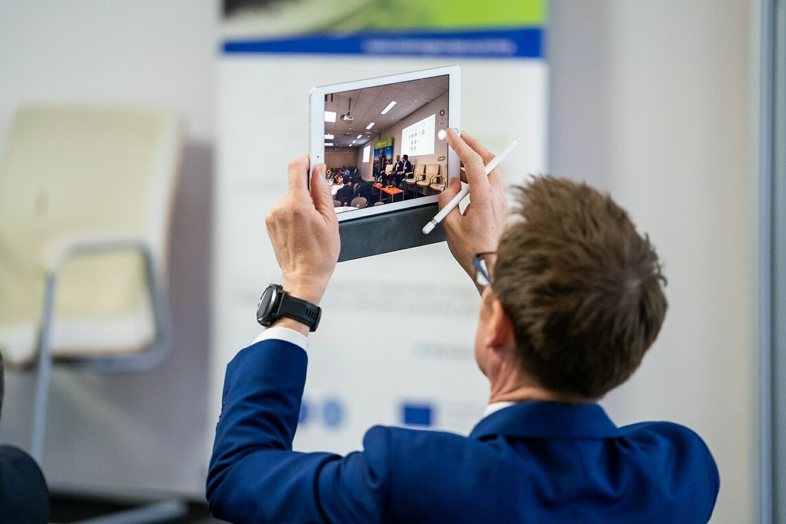 A man in a blue suit holds a white tablet and a stylus, taking a photo of a conference or presentation. A blurred screen and chairs are visible on the tablet's display. An empty chair is in the background