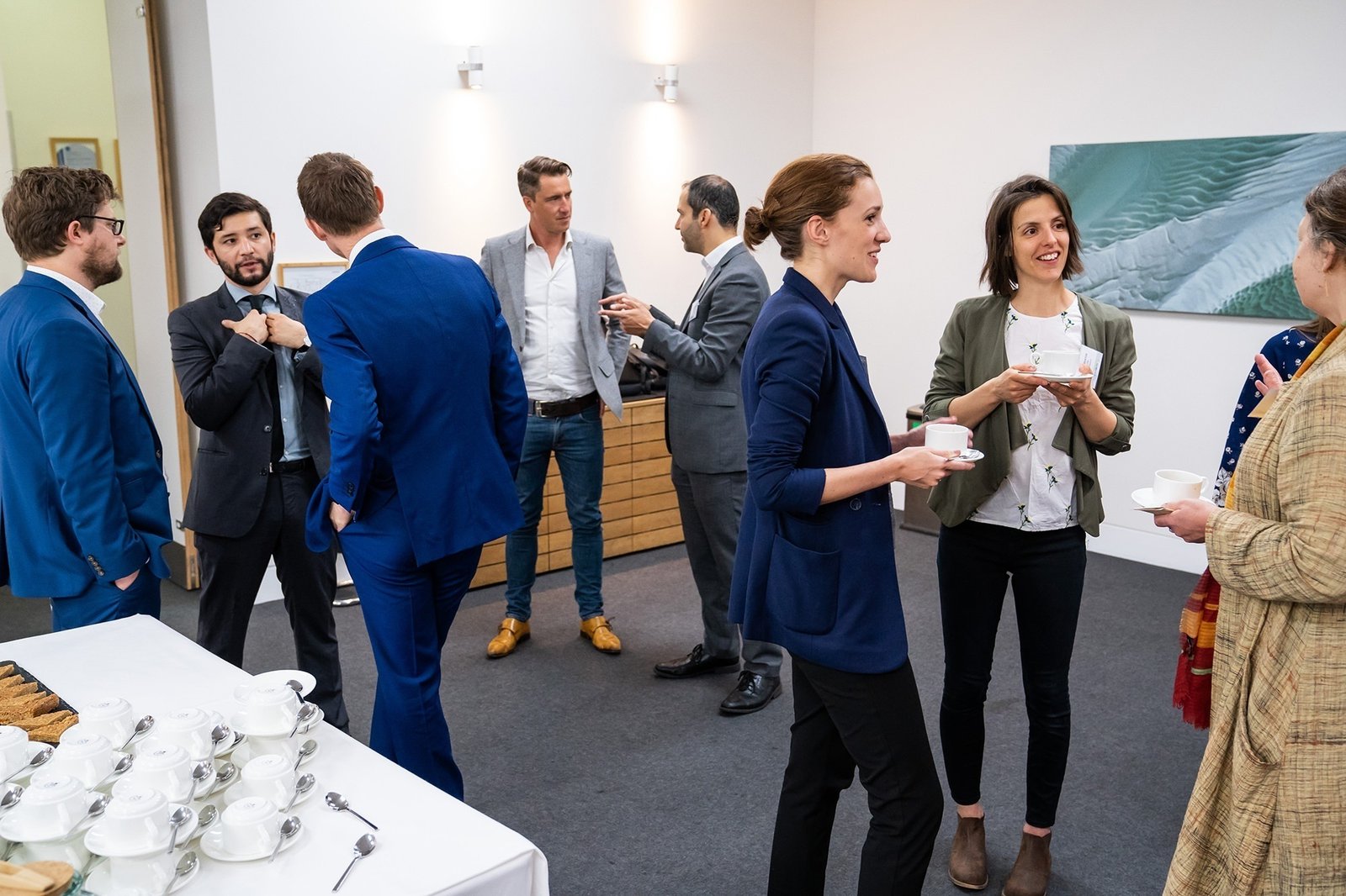 People in business attire stand and chat in a modern conference room. Some hold cups, engaging in conversation. There's a table with snacks and cups on the left. The room has white walls and a minimalist decor. James Gifford-Mead Photography - Event Photographer London