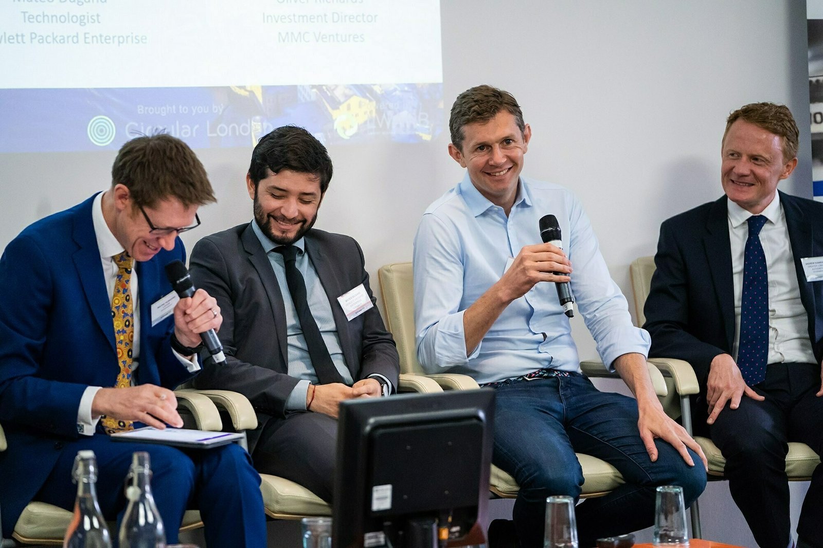 Four men in suits are sitting and smiling while participating in a panel discussion. One is holding a microphone and speaking. There are water bottles and glasses on the table in front of them. A projected presentation is visible in the background. James Gifford-Mead Photography - Event Photographer London