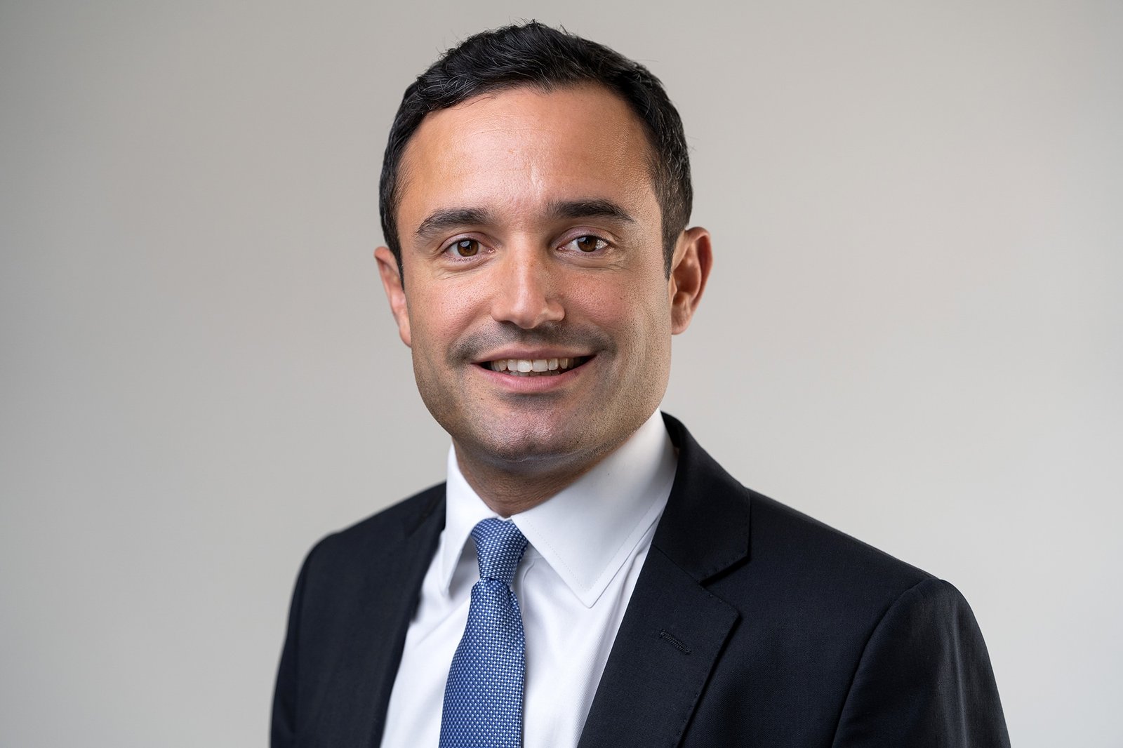 A man in a black suit and blue tie is smiling against a plain background. He has short, dark hair and is wearing a white shirt