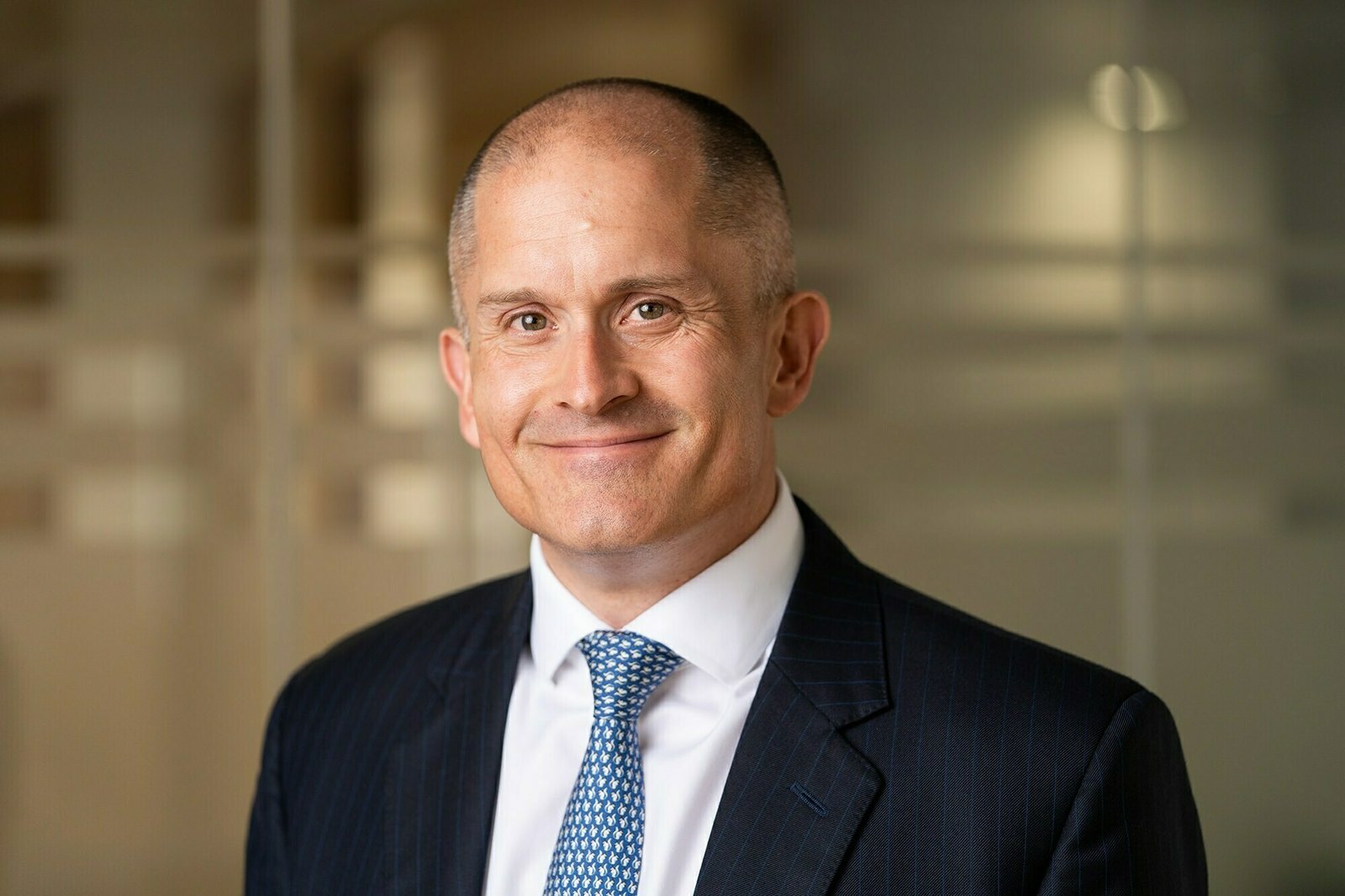 A bald man in a pinstripe suit and blue tie stands smiling. The blurred office background features glass walls with geometric patterns