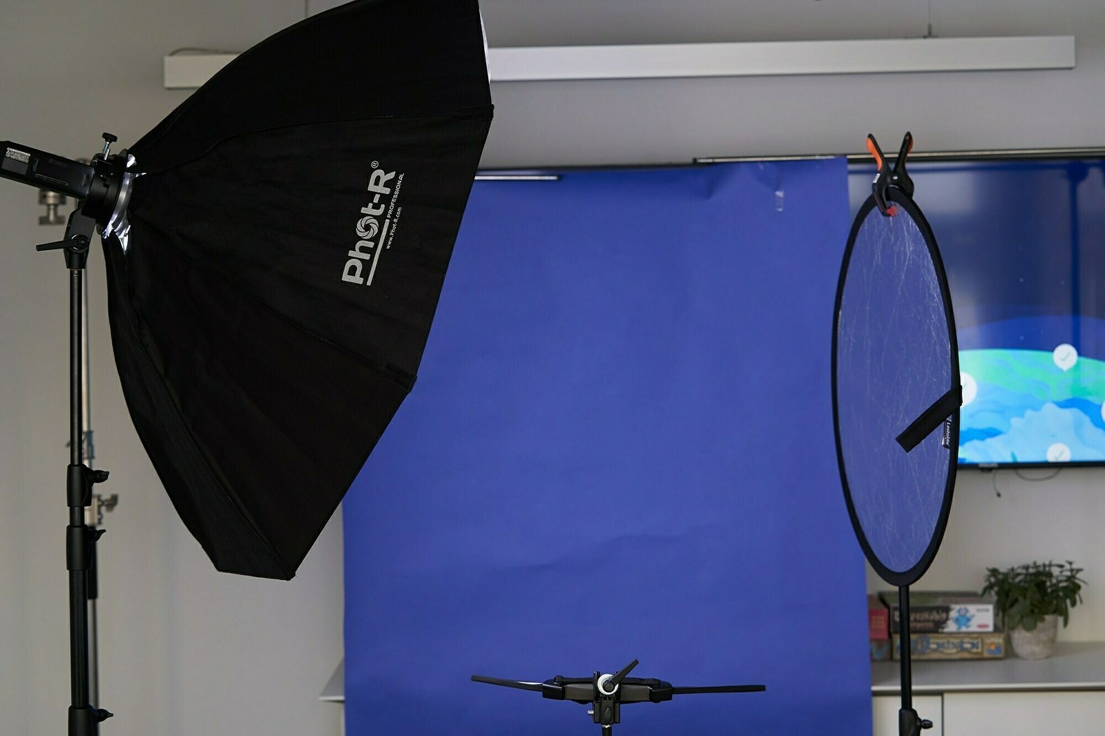 Photography studio setup with a black softbox light on the left, a circular reflector on the right, and a blue backdrop. A small camera or tripod base is visible in the foreground, with various studio equipment in the background. James Gifford-Mead Photography - Event Photographer London