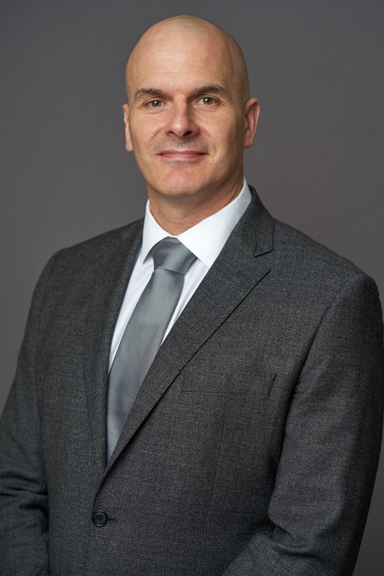 A man in a gray suit with a white shirt and gray tie stands against a plain gray background. He is bald and looking directly at the camera with a slight smile. James Gifford-Mead Photography - Event Photographer London