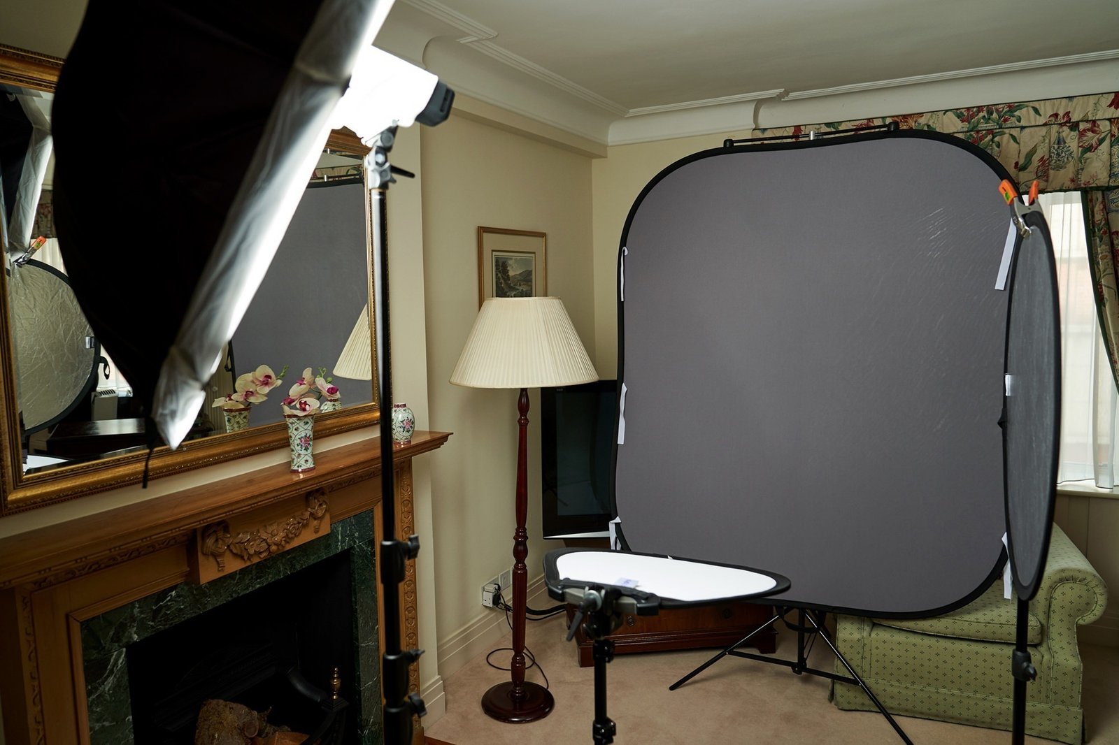 Room setup for photography with a large gray backdrop, studio lights, and a reflector. A lamp and floral curtains are visible, with a fireplace and mirror on the left side. James Gifford-Mead Photography - Event Photographer London