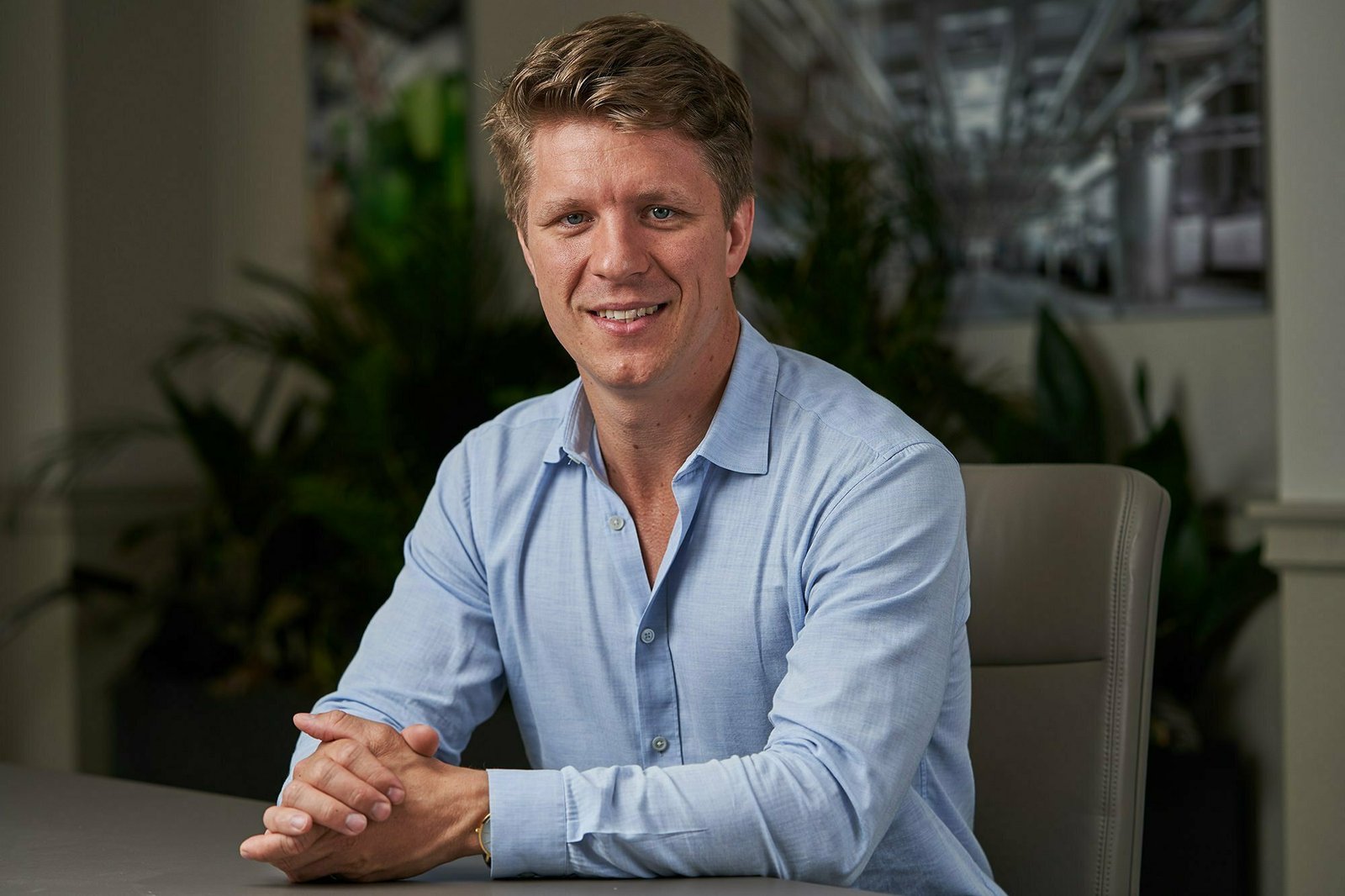 A person with short, light brown hair is sitting at a table, wearing a light blue button-up shirt. The background features blurred green plants and abstract art. The person has their hands clasped and is smiling at the camera