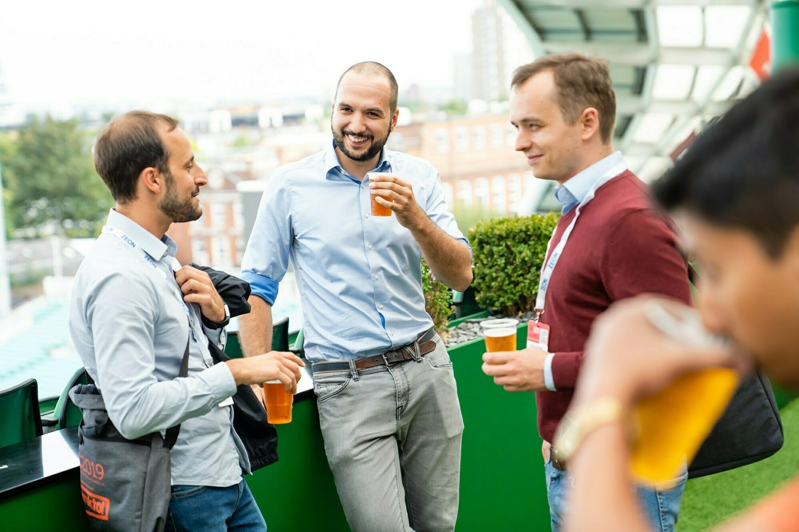 Four men stand on an outdoor balcony, holding beers and chatting. They appear relaxed and are wearing casual business attire. The background features city buildings and greenery