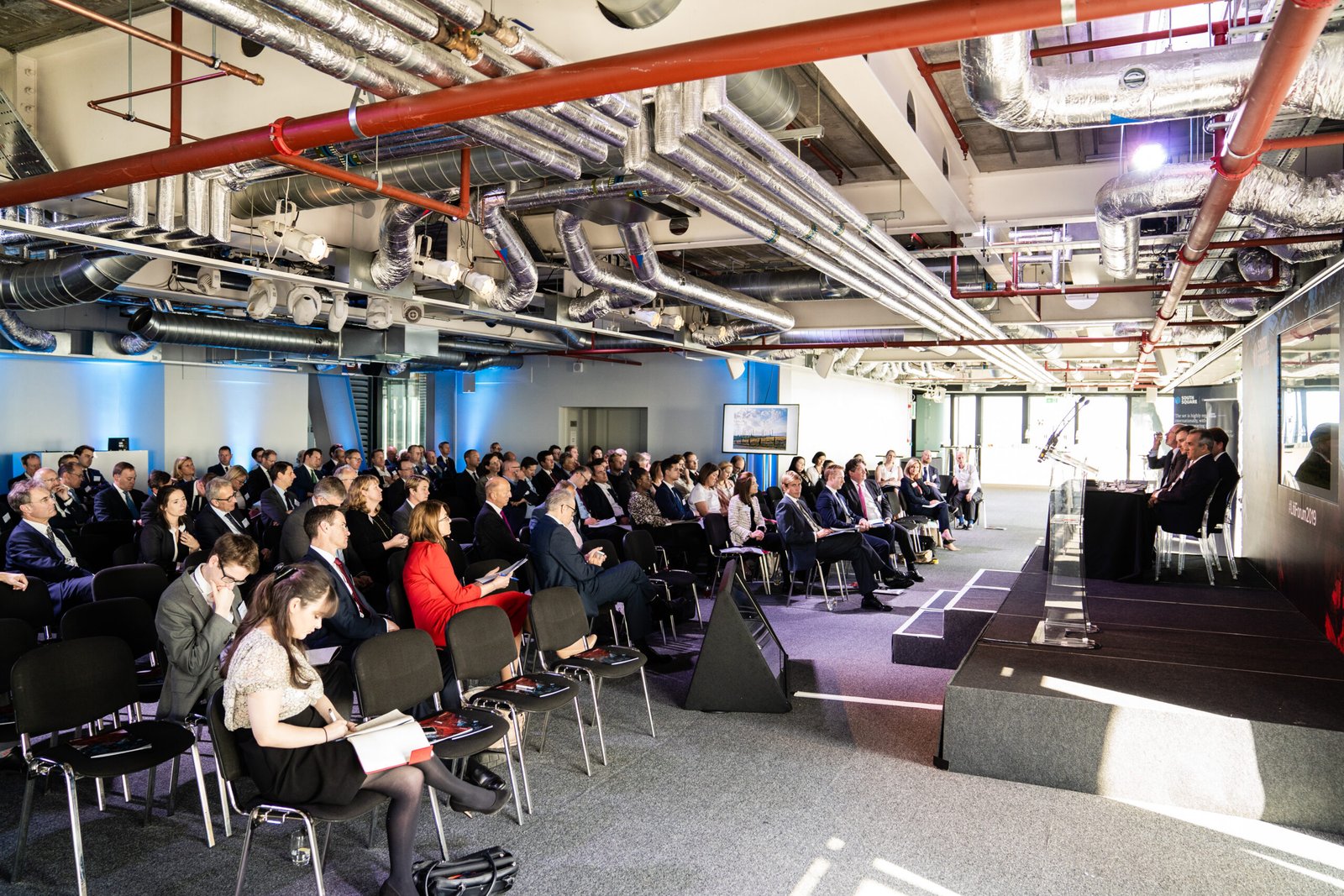 A large group of people seated in rows attending a conference in a modern industrial-style room with exposed pipes and ducts. Speakers are seated at a table in front, and attendees are focused on the presentation. James Gifford-Mead Photography - Event Photographer London
