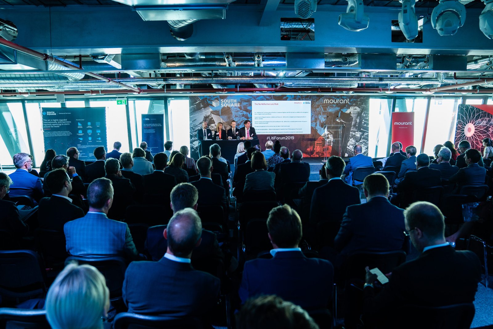 A packed conference room with attendees seated facing a panel of speakers at the front. The room has a modern design with a large screen displaying information behind the speakers. Cool lighting and reflections from ceilings and windows create a futuristic ambiance. James Gifford-Mead Photography - Event Photographer London