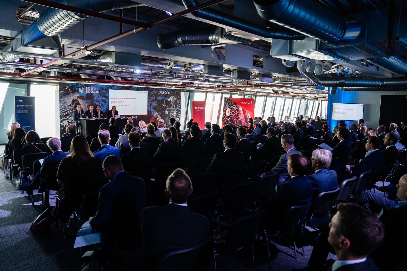 A large group of people in formal attire sit in rows facing a panel of speakers in a modern conference room with exposed ductwork. Presentation screens are visible at the front, and banners stand behind the presenters. James Gifford-Mead Photography - Event Photographer London