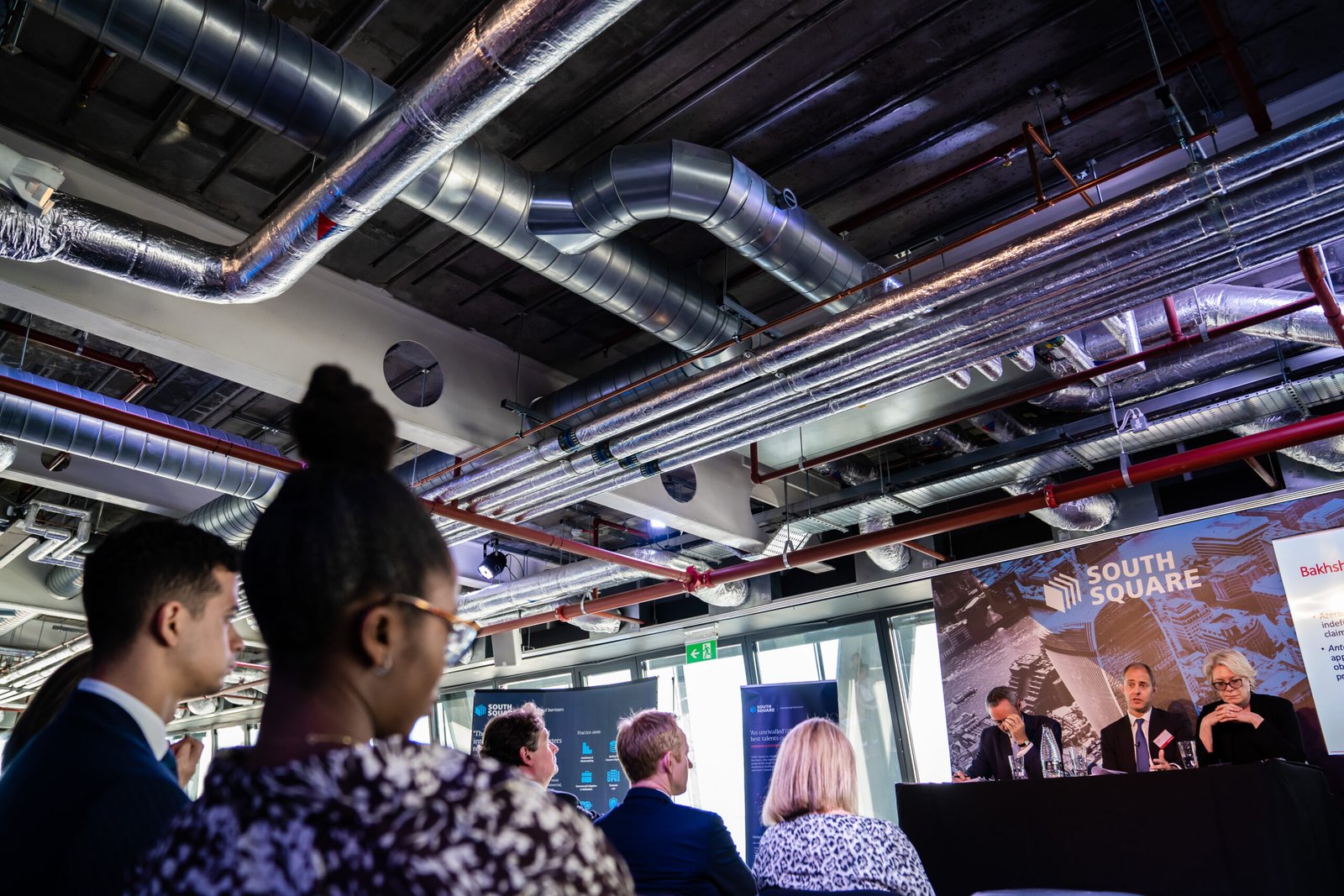 A group of people sit in an industrial-style room with exposed ducts, listening to a panel of three speakers seated on a stage. Banners behind the speakers read "South Square" and "Baker & Partners. James Gifford-Mead Photography - Event Photographer London