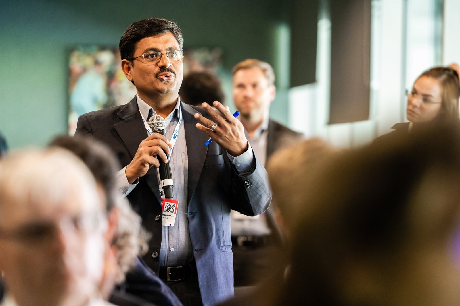 Man in a suit speaks into a microphone at a crowded event. Others in the background are listening attentively. The room has a professional setting, with people seated and some standing. James Gifford-Mead Photography - Event Photographer London