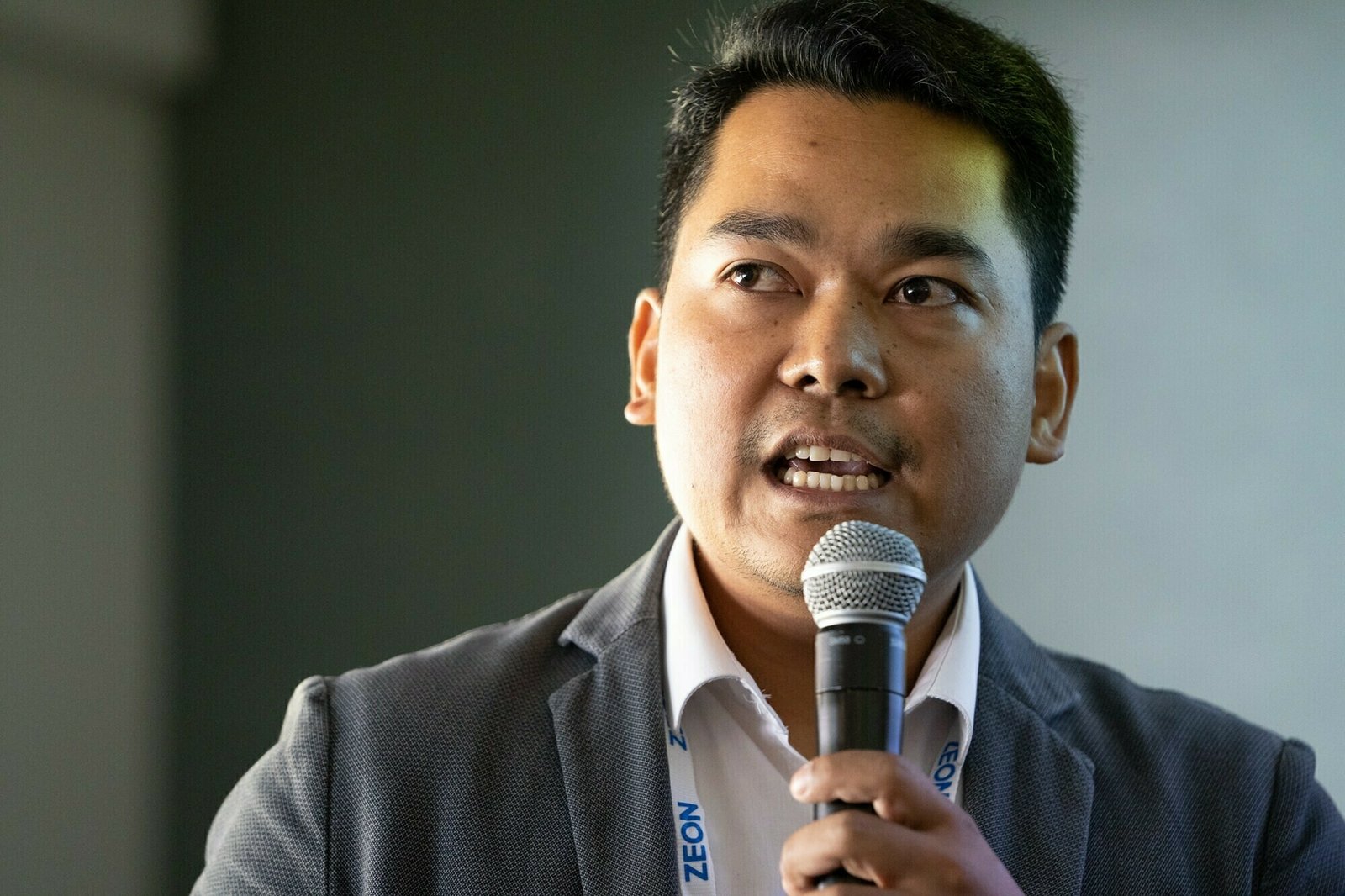 A man in a suit jacket holds a microphone while speaking. He wears a white shirt and appears engaged, looking slightly to the side. The background is blurred and neutral, focusing attention on the speaker. James Gifford-Mead Photography - Event Photographer London